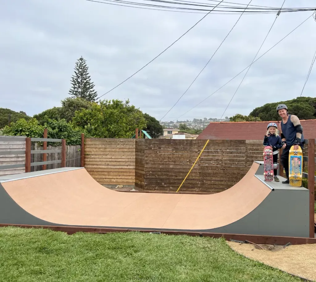 Three smiling children in pads and helmets with skateboards laying on flat bottom of 2x4 built miniramp