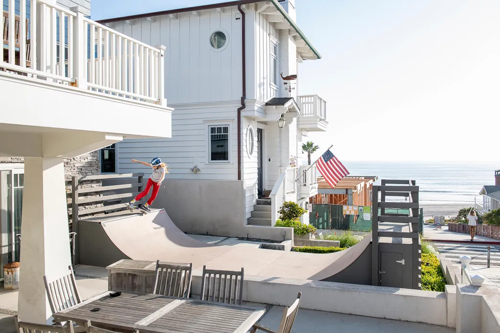 Child in red pants and helmet grinding backside grinding coping of 2x4 build miniramp in front yard of beach side
        home